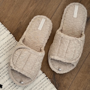 May include: A pair of beige open-toe slippers with a textured floral pattern. Each slipper has a wide strap across the top and a small label. The slippers are on a wooden surface next to a white and black patterned rug.