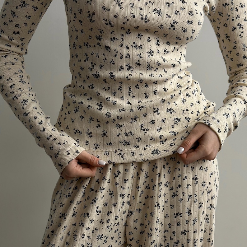 May include: Cream-colored ribbed knit long-sleeved top and matching pants set. The fabric is covered in a pattern of small, dark blue floral designs. The top has a high neckline and the pants appear to be a relaxed fit. The outfit is a comfortable loungewear set.