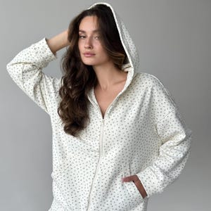 Op de afbeelding: Cr&egrave;mekleurige hoodie met ritssluiting en een delicaat bloemenpatroon. De hoodie heeft een capuchon, voorzakken en een ritssluiting over de hele lengte. De stof heeft een zachte, getextureerde uitstraling en het bloemmotief is in een gedempte groene kleur.