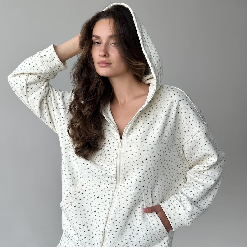 Op de afbeelding: Cr&egrave;mekleurige hoodie met ritssluiting en een delicaat bloemenpatroon. De hoodie heeft een capuchon, voorzakken en een ritssluiting over de hele lengte. De stof heeft een zachte, getextureerde uitstraling en het bloemmotief is in een gedempte groene kleur.