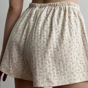 May include: Cream-colored shorts with a floral pattern. The shorts have a ribbed texture and an elastic waistband. Small pink and brown flowers are scattered across the fabric. The shorts appear to be made of a soft, comfortable material.