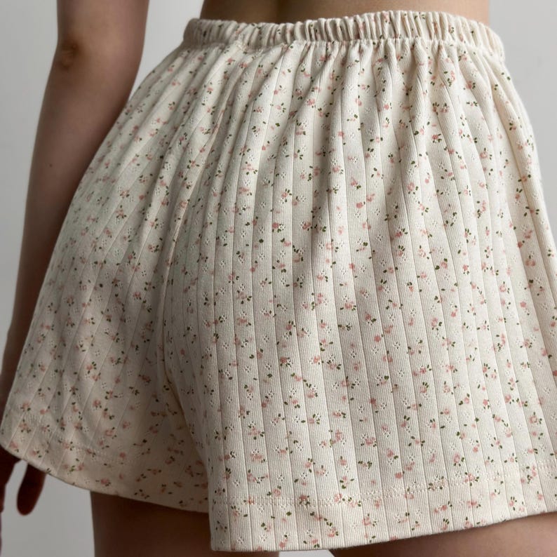May include: Cream-colored shorts with a floral pattern. The shorts have a ribbed texture and an elastic waistband. Small pink and brown flowers are scattered across the fabric. The shorts appear to be made of a soft, comfortable material.