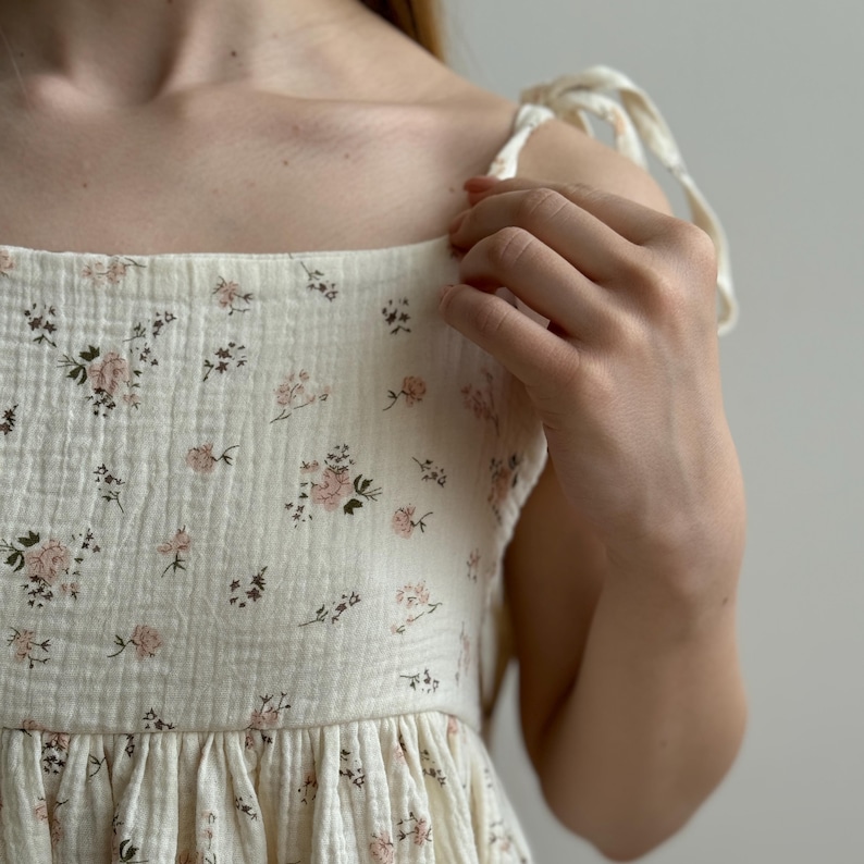 Op de afbeelding: Cr&egrave;mekleurige jurk met een bloemenpatroon met roze bloemen en groene bladeren. De jurk heeft strikbandjes en een vierkante halslijn. De stof lijkt een getextureerde katoen of mousseline te zijn.