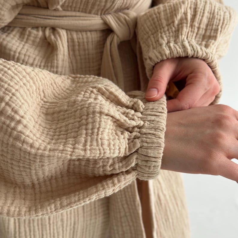 May include: Close-up of a beige robe with long sleeves. The sleeves are gathered at the wrist, creating a textured, ruched effect. The robe has a matching belt tied at the waist. The fabric appears soft and lightweight.