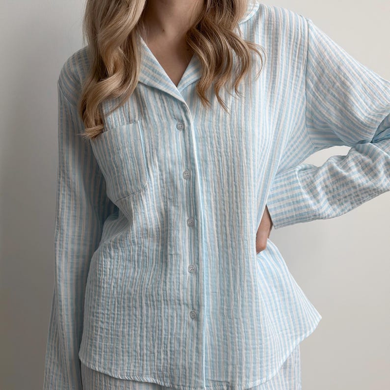 Op de afbeelding: Lichtblauw en wit gestreepte pyjama set. Het button-down shirt met lange mouwen heeft een borstzak en een klassieke kraag. De stof lijkt een zacht, getextureerd materiaal te zijn. De pyjama is een comfortabele nachtkleding.