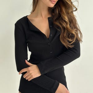 Pode incluir: Top preto de manga comprida com textura canelada e frente abotoada. O top tem decote em forma de barco e é feito de um material macio e justo. As mangas são compridas e justas, e o design geral é simples e elegante.