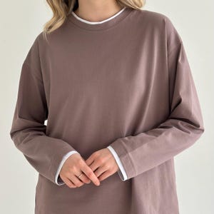 Pode incluir: Uma camisa de manga comprida em tom malva suave com um design em camadas, apresentando uma gola redonda branca e punhos brancos visíveis nos pulsos. A peça tem um corte descontraído com ombros descaídos e um acabamento de tecido liso.