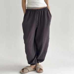 May include: Dark grey, loose-fitting pants with a drawstring waist and elastic cuffs. The pants have a textured, crinkled appearance. Beige cross-strap sandals are worn with the pants. The person is wearing a white tank top.