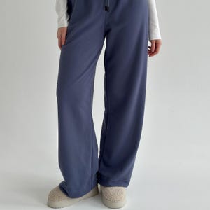 May include: A pair of wide-leg, slate blue sweatpants with a drawstring waist. The pants are paired with a white long-sleeve shirt and beige slippers. The casual pants are made of a soft material.