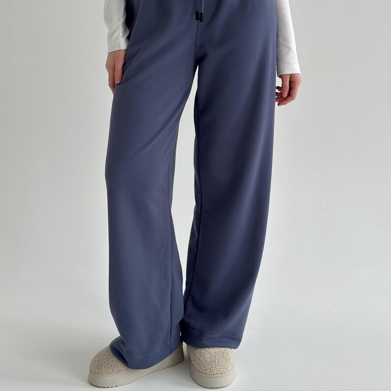 May include: A pair of wide-leg, slate blue sweatpants with a drawstring waist. The pants are paired with a white long-sleeve shirt and beige slippers. The casual pants are made of a soft material.