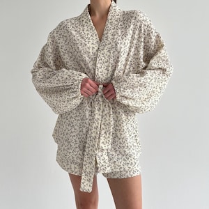 Op de afbeelding: Crèmekleurige robe en short set met bloemenprint. De robe heeft een V-hals, lange, volumineuze mouwen en een bijpassende strikceintuur. De shorts zijn hoog getailleerd en hebben een vergelijkbaar bloemenpatroon. De stof lijkt zacht en licht.