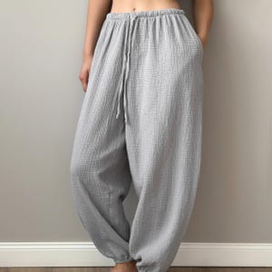 May include: Light gray harem pants with a drawstring waist and pockets. The pants are made of a textured, lightweight fabric. The loose fit and gathered ankles create a relaxed silhouette. These pants are a comfortable and stylish choice for casual wear.
