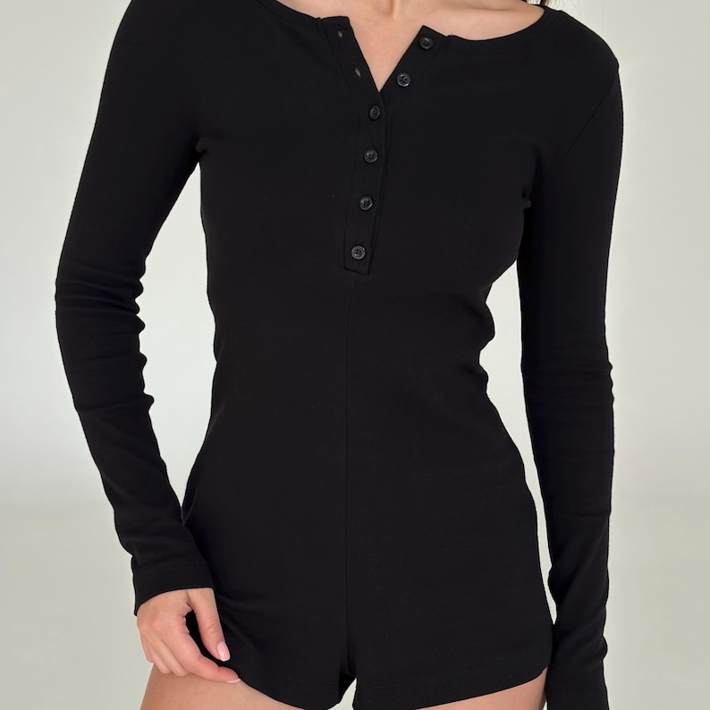 May include: Black long-sleeved romper with a scoop neckline and button-down front. The one-piece garment has a fitted shape and short shorts. The fabric looks like ribbed knit, offering a comfortable and stylish appearance.