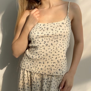 May include: A cream-coloured camisole and skirt set featuring a small, dark floral pattern. The camisole has thin straps and a scoop neckline. The skirt has a matching floral print and a relaxed fit. The fabric appears to be a soft, ribbed knit.