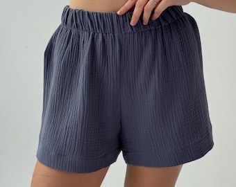 May include: Dark slate blue shorts featuring a textured, crinkled fabric finish. These shorts have a high-rise elastic waistband with gathered detailing and a relaxed fit through the legs, ending in a simple hemline with visible stitching.