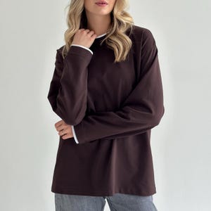 Pode incluir: Uma camisa de manga comprida castanho escuro com acabamento branco na gola e punhos. A camisa é feita de um material macio e tem um corte folgado. A modelo está a usar a camisa com calças de ganga cinzentas.
