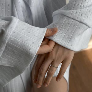 May include: A silver ring with a thin band, adorned with small, sparkling stones. The ring is worn on a hand, with a white, textured long-sleeve shirt in the background. The shirt has a subtle pattern of small, evenly spaced holes.