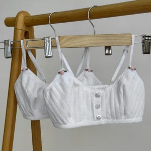 Peut inclure: Deux brassières blanches côtelées à fines bretelles réglables, chacune ornée d'une petite rose rose à la base de la bretelle. L'avant des brassières présente une rangée verticale de trois boutons blancs et un ourlet festonné.