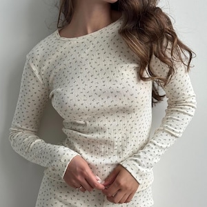 May include: A long-sleeved, ribbed knit top in an off-white colour featuring a small, repeating dark floral pattern. The garment has a round neckline and a fitted silhouette, with the fabric texture showing vertical lines throughout the design.