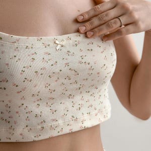 Op de afbeelding: Cr&egrave;mekleurige tube top met een delicaat bloemenpatroon in roze en groen. Een kleine strik is gecentreerd op de top. De top is gemaakt van een zachte, geribbelde stof.