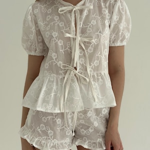 May include: White embroidered two-piece set featuring a top with short, puffed sleeves and a tie-up front. The matching shorts have a ruffled hem. The set is adorned with a delicate floral pattern, creating a light and airy aesthetic.