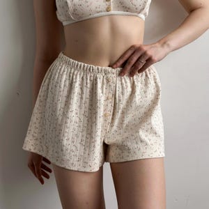 May include: Cream-colored pajama shorts and matching top with a floral pattern. The shorts have an elastic waistband and button closure. The top is a bralette style with button details. The fabric has a ribbed texture.
