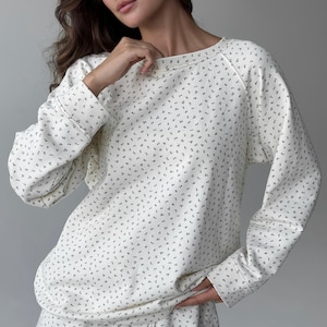 May include: Cream-colored long-sleeved sweatshirt with a small, repeating floral pattern in a darker shade. The sweatshirt has a crew neck and ribbed cuffs. The fabric appears soft and comfortable, suitable for casual wear.