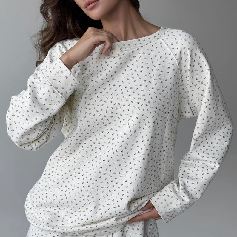 May include: Cream-colored long-sleeved sweatshirt with a small, repeating floral pattern in a darker shade. The sweatshirt has a crew neck and ribbed cuffs. The fabric appears soft and comfortable, suitable for casual wear.