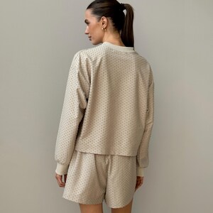 Puede incluir: Conjunto de sal&oacute;n de dos piezas beige. El conjunto incluye una parte superior de manga larga y pantalones cortos a juego. La parte superior tiene cuello redondo y los pantalones cortos tienen cintura con cord&oacute;n. La tela tiene un patr&oacute;n texturizado y punteado. Los pu&ntilde;os y el cuello son de color beige claro.