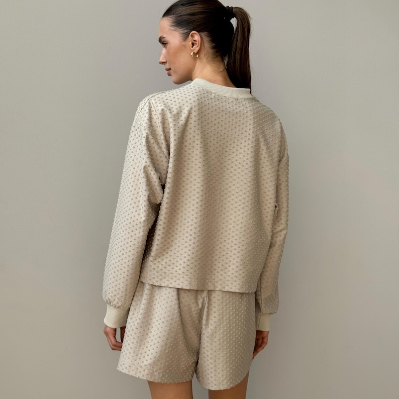 Puede incluir: Conjunto de sal&oacute;n de dos piezas beige. El conjunto incluye una parte superior de manga larga y pantalones cortos a juego. La parte superior tiene cuello redondo y los pantalones cortos tienen cintura con cord&oacute;n. La tela tiene un patr&oacute;n texturizado y punteado. Los pu&ntilde;os y el cuello son de color beige claro.