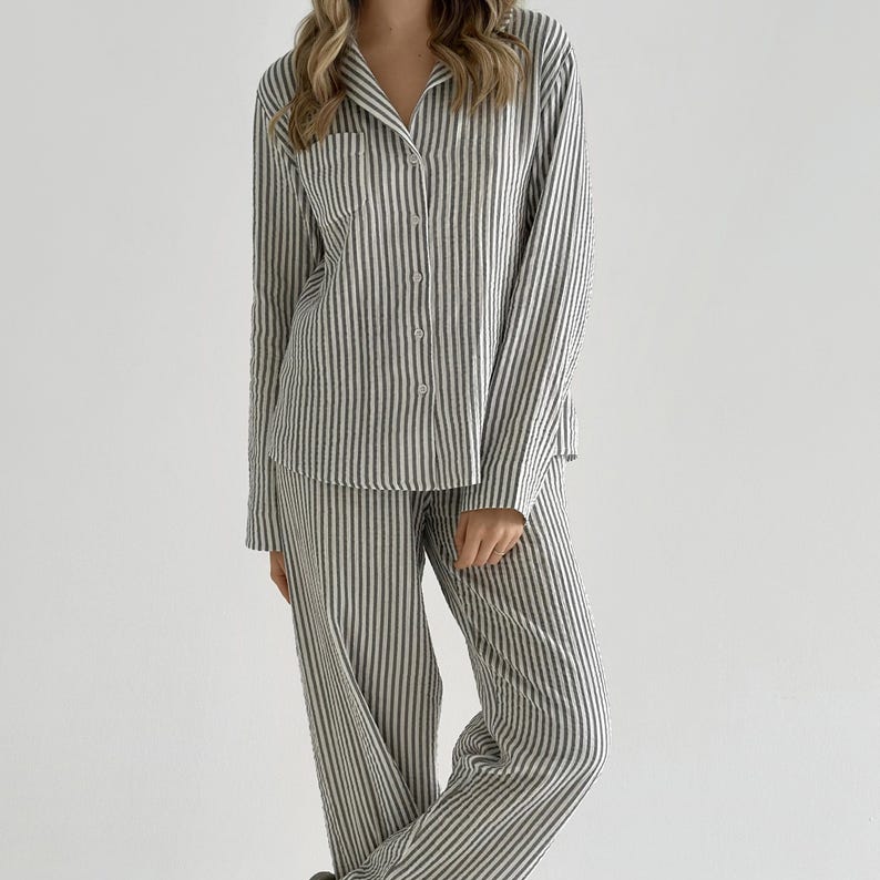 Op de afbeelding: Een tweedelige pyjamaset met een verticaal streepjespatroon in grijstinten en wit. Het overhemd met lange mouwen en knoopsluiting heeft een borstzak. De bijpassende broek heeft een relaxte pasvorm. De pyjama is gemaakt van een zachte, lichte stof.