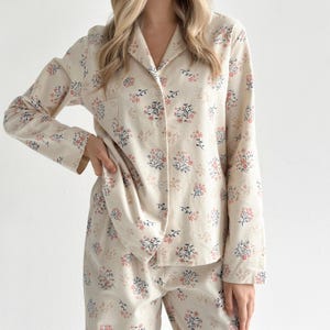 Könnte beinhalten: Ein langärmeliges Schlafanzug-Set bestehend aus einem geknöpften Oberteil und einer passenden Hose aus cremefarbenem Stoff. Das Kleidungsstück zeigt ein florales Muster mit kleinen rosa Blüten, dunkelblauen Blättern und dünnen Stielen. Das Oberteil hat einen Reverskragen.