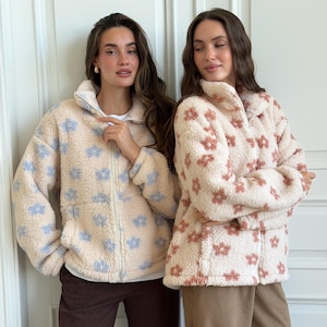 Könnte beinhalten: Zwei Fleecejacken mit Reißverschluss, Stehkragen und langen Ärmeln. Eine Jacke ist cremefarben mit hellblauem Blumenmuster, die andere ist cremefarben mit terrakottafarbenem Blumenmuster. Beide verfügen über einen Reißverschluss vorne.