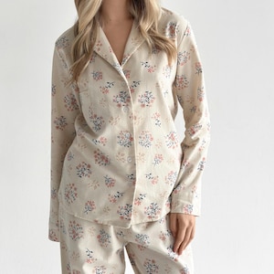 Puede incluir: Conjunto de pijama color crema con estampado floral. El conjunto incluye una camisa abotonada con cuello y un pantalón a juego. El diseño floral presenta flores rosas y azules con hojas verdes. El pijama está hecho de una tela suave y cómoda.