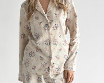 Puede incluir: Un conjunto de pijama de manga larga con botones, compuesto por una camisa y pantalones de tela color crema con un estampado floral repetitivo de pequeñas flores rosas y tallos de hojas azul oscuro. La camisa tiene cuello de solapa y bolsillo.