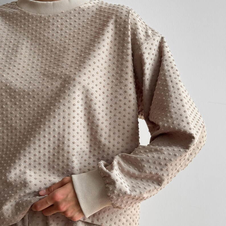 Puede incluir: Sudadera beige con un patr&oacute;n texturizado de puntos. La parte superior de manga larga tiene cuello redondo y pu&ntilde;os acanalados. La tela parece suave y c&oacute;moda, adecuada para ropa casual.