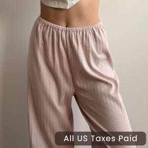 May include: Pale pink, wide-leg pants with an elastic waistband. The pants have a subtle, vertical ribbed texture with small, diamond-shaped perforations. The text "ALL US Taxes Paid" is at the bottom of the image. The pants appear to be made of a soft, lightweight fabric.