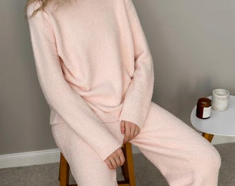 May include: A light pink, two-piece lounge set. The set includes a crew neck sweater and matching pants. The fabric has a soft, textured appearance. The person is sitting on a wooden stool.