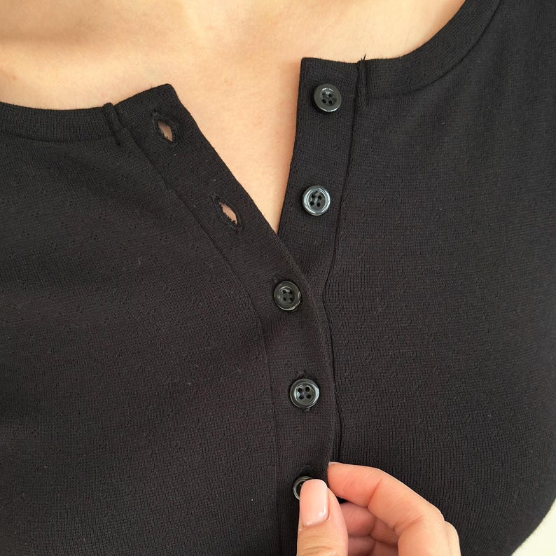 May include: Close-up of a black henley shirt with a scoop neck and a button-down placket. The shirt has four black buttons and a textured fabric. A hand is buttoning the shirt.