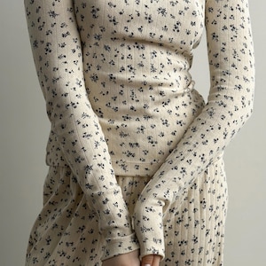 May include: Cream-colored ribbed long-sleeved top and matching pants set. The fabric is covered in a delicate pattern of small, dark blue floral designs. The outfit has a comfortable, relaxed fit, suitable for loungewear or casual wear.