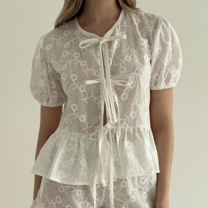 May include: White embroidered top with short, puffed sleeves and a tie closure. The top features a floral pattern and a ruffled hem. The fabric appears sheer, and the design is delicate and feminine.