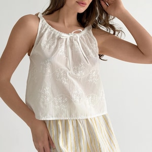 May include: A white sleeveless top featuring an eyelet floral pattern, a gathered neckline with a thin ribbon tie, and a loose fit. Below the top is a garment with vertical yellow and white stripes, completing the two-piece set.