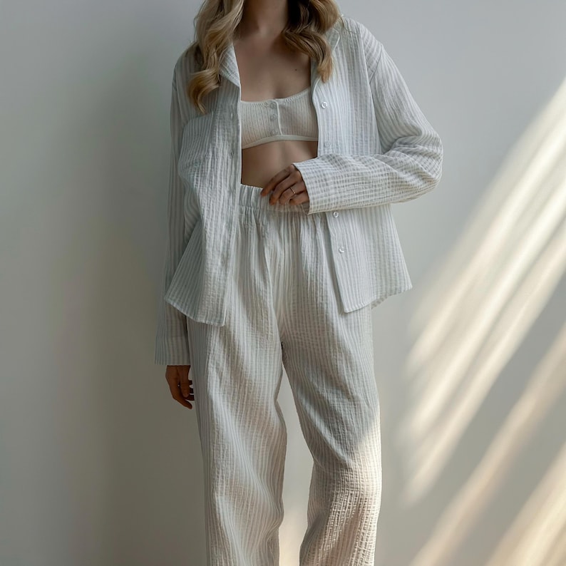 Op de afbeelding: Een driedelige witte gestreepte pyjamaset. De set bevat een overhemd met knopen, een bralette en een bijpassende broek. De stof heeft een getextureerde uitstraling. De outfit is ontworpen voor comfort en ontspanning.