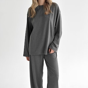 May include: A charcoal gray lounge set consisting of a long-sleeved crew neck top and matching wide-leg trousers. The garment features a solid, smooth fabric texture with a relaxed fit, long sleeves, and a simple, unadorned design throughout.