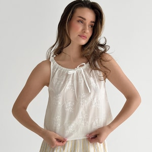 May include: A sleeveless white top featuring a delicate eyelet floral pattern and a gathered neckline with a thin white ribbon tie. The fabric has a subtle texture with repeating embroidered wave motifs across the front of the garment.