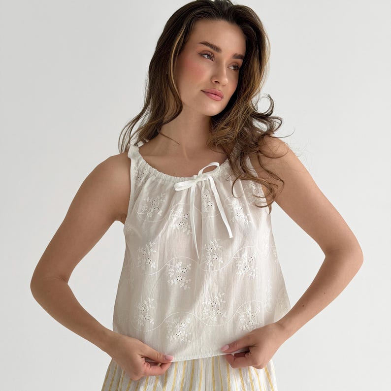May include: A sleeveless white top featuring a delicate eyelet floral pattern and a gathered neckline with a thin white ribbon tie. The fabric has a subtle texture with repeating embroidered wave motifs across the front of the garment.