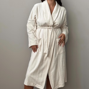 May include: Cream-colored, long-sleeved robe with a shawl collar and a matching belt. The robe has two front pockets and a vertical striped pattern. The robe is made of a soft, textured fabric.