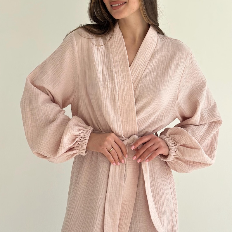 May include: A light pink robe with long sleeves and a tie waist. The robe has a textured fabric and a relaxed fit. The sleeves are gathered at the wrists, and the robe is open in the front.