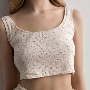 Può includere: Top corto a coste color crema con un delicato motivo floreale rosa e marrone. Il top presenta uno scollo tondo e un piccolo dettaglio a fiocco al centro. Il tessuto ha un design a coste verticali strutturato.