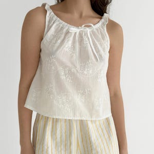 May include: A white sleeveless top featuring an eyelet lace floral pattern with wavy embroidered lines and a drawstring neckline with a tie closure. The top is paired with yellow and white vertical striped shorts with a textured fabric finish.